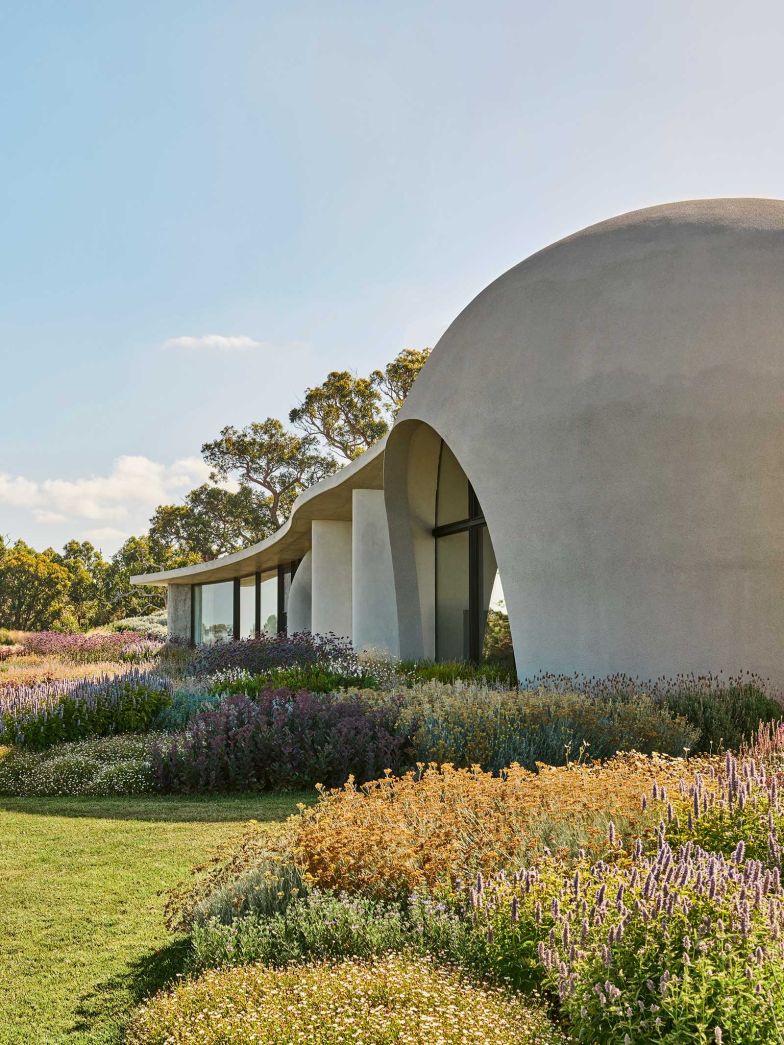 Ein dichter Gräsergarten legt sich wie ein weiches Band um das organisch geformte Haus und nimmt den Ton der Landschaft auf. Seine wechselnden Farben bringen die rohe Betonfassade sanft zum Leuchten.