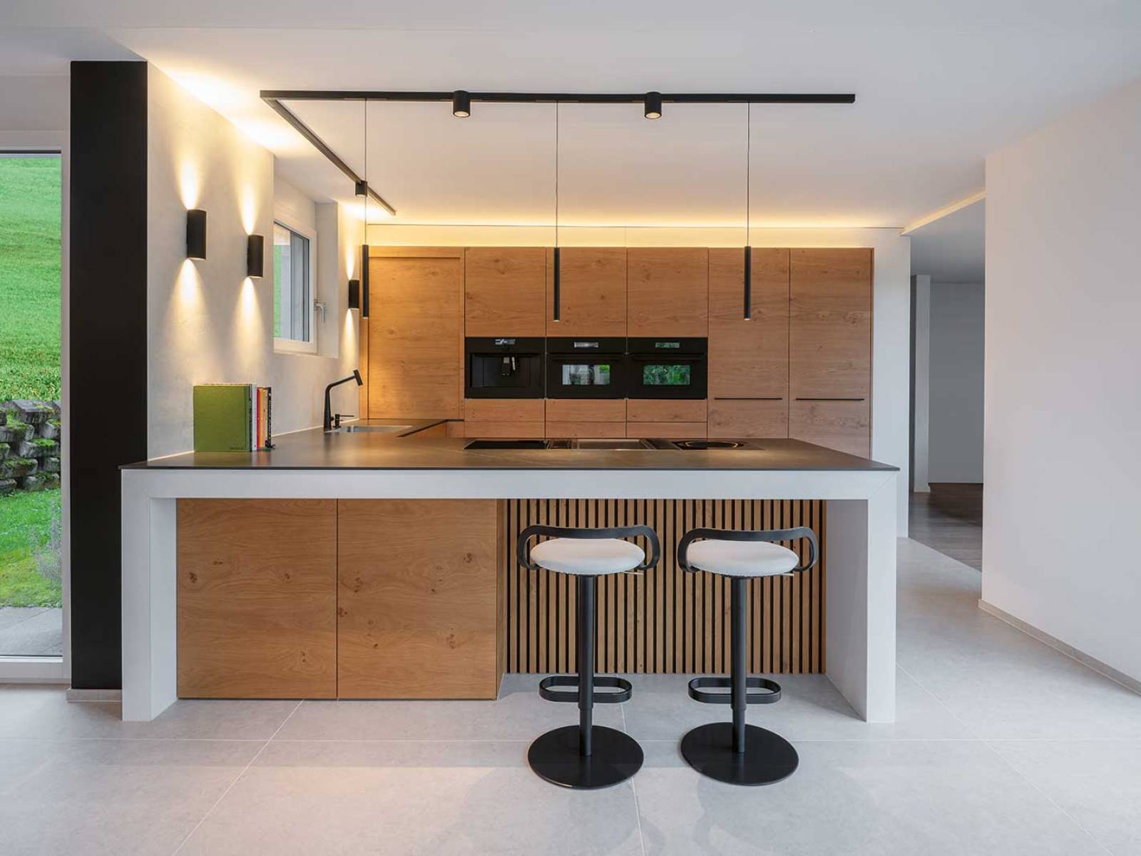In warmer Eiche und mit klaren Linien präsentiert sich diese offene Küche. Die weisse Insel mit feiner Holzverkleidung und zwei bequemen Barhockern bildet ein einladendes Zentrum. Präzise schwarze Akzente – Abdeckung, Griffe, Armaturen, Spotlights – verleihen Struktur, während das Lichtkonzept Tiefe und Funktionalität schafft. ELBAU.